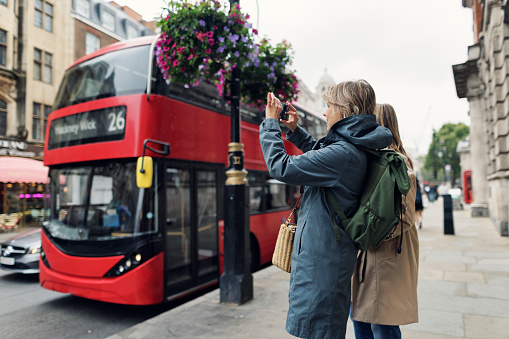 Mother and teenage daughter sightseeing London, UK. Overcast summer day. Mother is taking photo of a red double decker bus.\nCanon R5