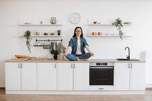 Peaceful asian woman meditating while sitting on white designer kitchen set. Slender female dressed in casual clothes practicing yoga to achive life balance and harmony at cozy apartment.