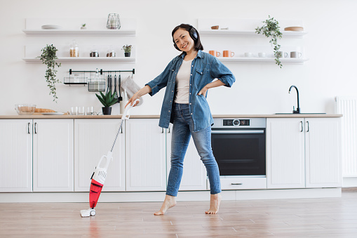 Charming korean lady without footwear standing on tiptoes with lightweight stick vacuum in hand at home. Joyful slim woman dancing for melody in headphones while dusting off bare floor in kitchen.