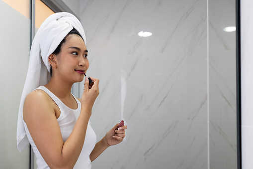 A young Asian Woman getting ready for work doing a morning makeup routine in the bathroom mirror at home. paint brush on cheeks, apply lip, and dry hair with a towel