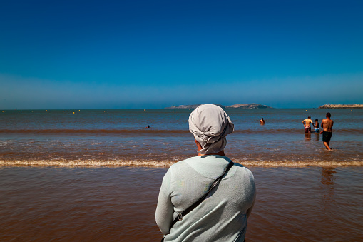 Essaouira, Marocco - Agosto 3, 2023: A woman in traditional clothing and a headdress watches the sea from the beach. The Muslim religion prevents women from showing their bare bodies on mixed beaches.