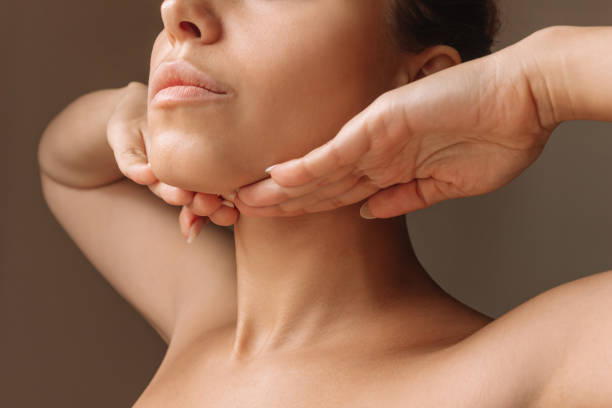 Young woman touching under the chin with hands massaging her face. Exercises from the second chin Cropped shot of young caucasian woman touching under the chin with hands massaging her face on dark brown background. Rejuvenation, facelift, facefitness. Exercises from the second chin, pelican neck chin stock pictures, royalty-free photos & images