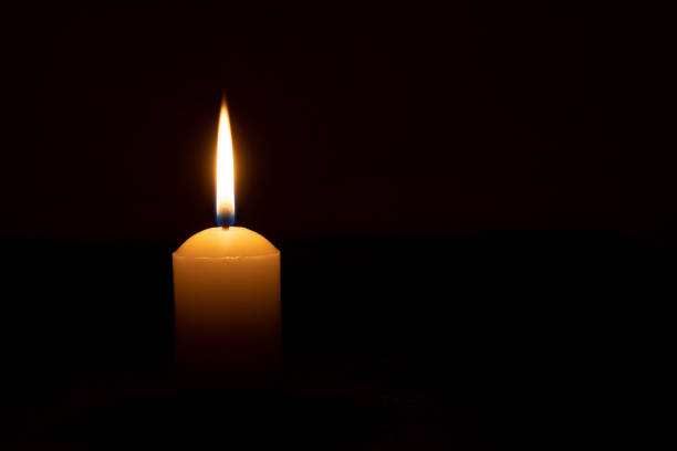 istock Single burning candle flame or light glowing on a big white candle on black or dark background on table in church for Christmas, funeral or memorial service with copy space. 1642422013