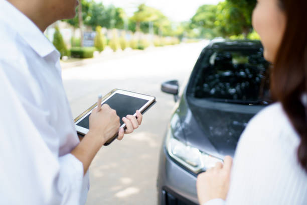 Land vehicle accident, an insurance claim agent - officer inspecting the damaged car at the front bumper and write down or note an inspecting report and taking a photo on the digital tablet. Woman discussing with a claims agent about her damaged car.