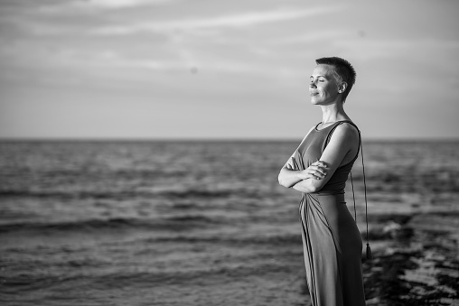A relaxed woman with a short haircut in a dress stands on the ocean shore with her face towards the setting sun. A middle-aged woman with a slight smile.
