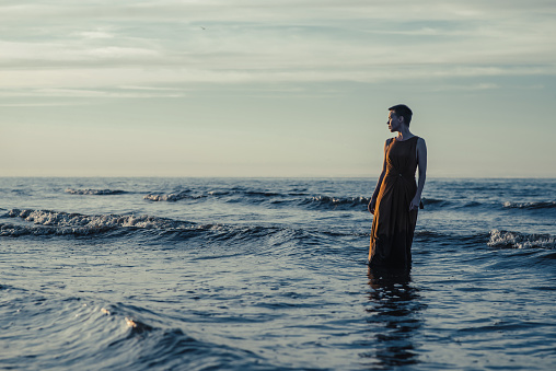 A woman is standing in a long dress in the water in the ocean or sea. A woman with short hair looks at the sunset. Reflections, crisis.