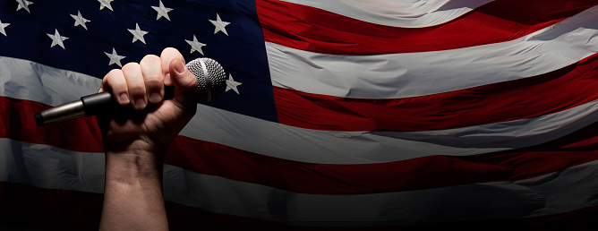 Male Fist Gripping Microphone Over Waving American Flag Background Banner.