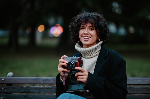Cropped shot of a joyful young pretty girl sitting on a bench at the park and holding her old retro camera in her hands while looking straight into the camera with a big smile on her face. Copy space.