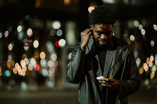 Portrait of a young urban African American man standing on the city street downtown at night and putting earbuds on. A young urban man is using cutting-edge technologies on the street.