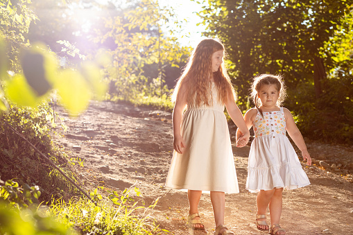 Two little girls, sisters walking outdoors in white dresses, holding hands. Copy space