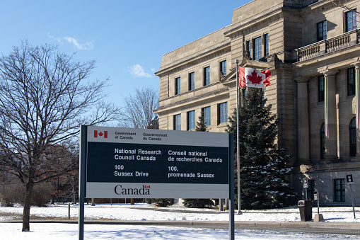 A sign out front of the National Research Council of Canada government office building is seen with the Canadian flag waving in the background.
