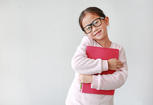 Asian schoolgirl wearing eyeglass hug a book and holding pencil in hand against white background with copy space. Portraits of child girl looking straight at camera.