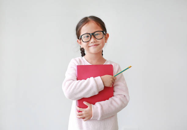 estudante asiática usando óculos abraçar um livro e segurando lápis na mão contra fundo branco. retratos de menina criança olhando diretamente para a câmera. - child glasses elementary student reading - fotografias e filmes do acervo