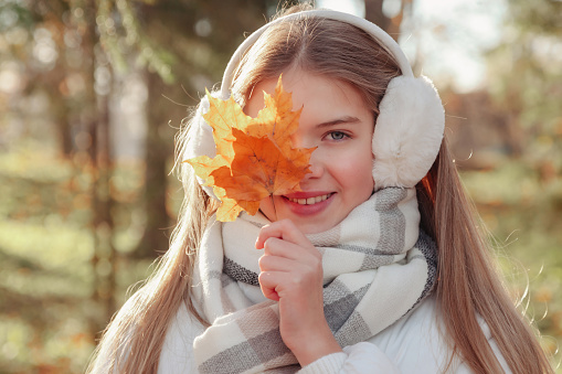 Face of lovely teen cover girl covering eye with fall leaves in autumn park, smile looking at camera. Cute young lady model in white jacket in fall forest. Leisure activity concept. Copy ad text space