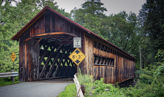 Covered bridge stretches over a river or stream. Made of wood planks and lattice work. A weight limit sign on the road and green trees in the background. Late summer in New Hampshire.