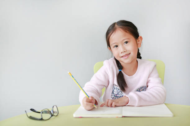 petite fille asiatique souriante écrit dans un livre ou un cahier avec un crayon sur la table de la classe sur fond blanc. portraits d’enfants regardant directement la caméra. - schoolgirl little girls crayon human face photos et images de collection