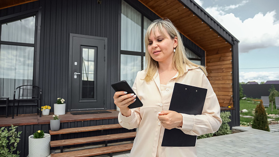 Real estate agent sending location of cottage residence in suburban area to potential clients via phone. real estate agent stands past house offered by company