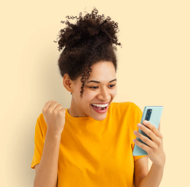 Excited happy woman holding mobile phone Female reading good news online shopping Excited happy young African American woman holding mobile phone and looking at screen gesturing yes with clenched fist. Female reading good news using smartphone, online shopping concept excited woman yellow stock pictures, royalty-free photos & images