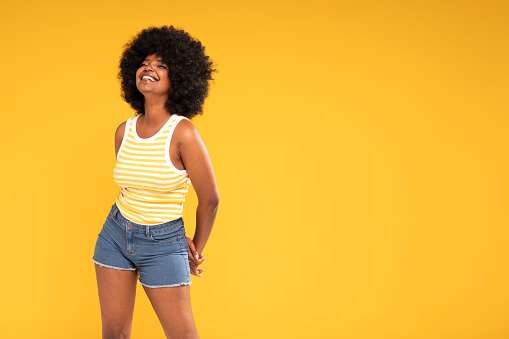 Photo of charismatic cute african american girl in trendy jeans shorts and top laughing happily. Vibrant yellow studio background. A lot of copy space. Charming woman with afro hairstyle.Real people emotions.