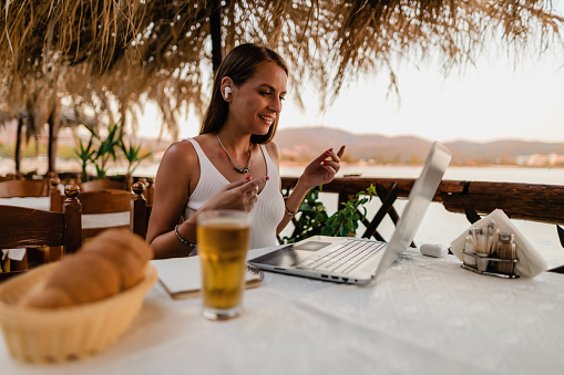 Beautiful young woman sitting in a restaurant at the seaside and using a laptop during her summer vacation in Greece