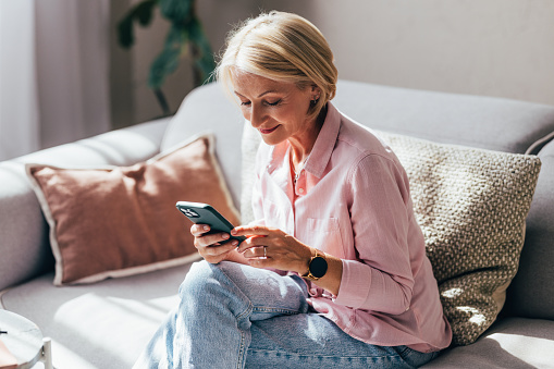A smiling Caucasian female using her smartphone while relaxing on the cozy sofa at home.