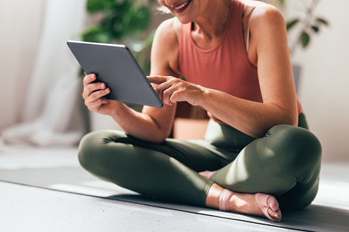A wide view of a smiling Caucasian female using her tablet while sitting on exercise mat at home.