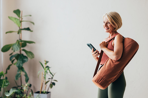 A side view of a smiling Caucasian female (carrying a yoga mat in a bag) using her smartphone while standing at home.