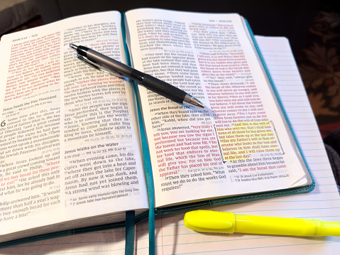 Open bible sitting on notebook paper. Ballpoint pen pointing to highlighted passage in John 6:39. Highlight is bright yellow. Teal bible bookmark ribbon down center of Bible. Teal edges of bible leather showing. No writing on notebook page. Yellow highlighter is slightly visible in bottom left corner