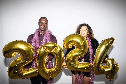 A men and a women standing and smiling at the camera while holding inflated gold numbers to welcome the New Year 2024 against a white background