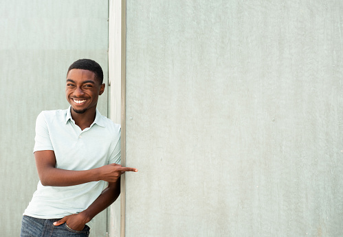 Portrait happy young African man leaning against wall and pointing finger to empty space