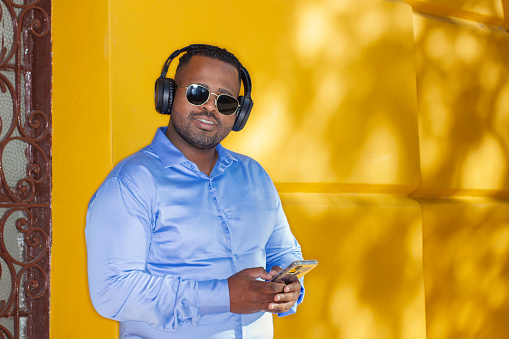 Portrait of young man with sunglasses and wireless heaphones leaning against yelow wall