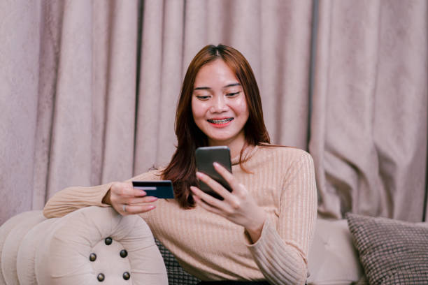 istock Beautiful asian woman sitting on couch while online purchasing using smartphone. 1643291498