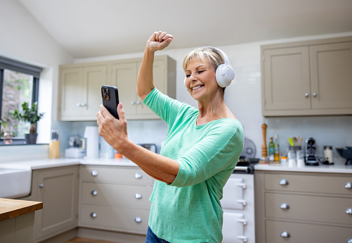 Senior woman looking very happy dancing at home while listening to music on her cell phone using headphones - retirement concepts
