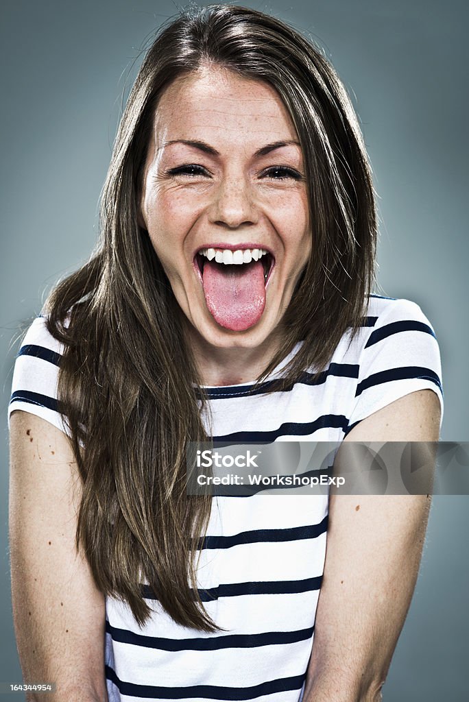 Joyous young woman in striped top sticking out her tongue Young Happy Woman Sticking Out Her Tongue Over a Grey Background Young Women Stock Photo