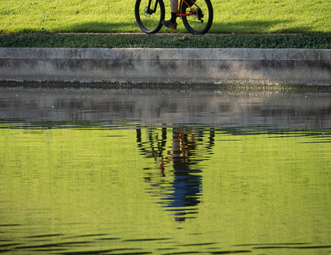 The Woodlands, TX, USA - August 27, 2023: Male bicyclist riding on a sidewalk on the riverbank with his rippled reflection in the water.