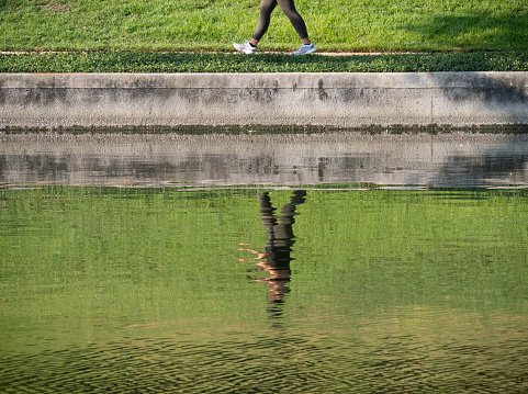 The Woodlands, TX, USA - August 27, 2023: Woman walking along a riverbank sidewalk with her rippled reflection captured in the photograph.