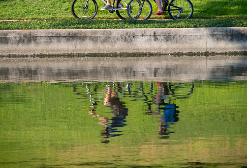 The Woodlands, TX, USA - August 27, 2023: Pair of bicyclists riding on a sidewalk on the riverbank with their rippled reflections in the water.