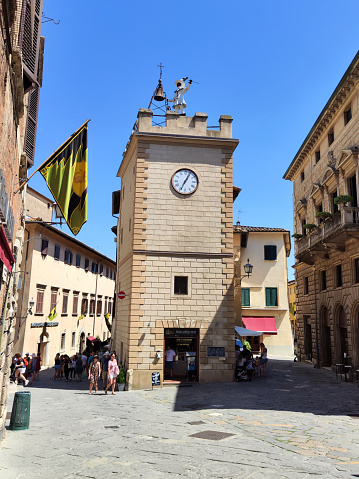Tourists walking on the street by the famous tower of Pulcinella, a landmark clock tower in Montepulciano,  a medieval and Renaissance hill town in the province of Siena in southern Tuscany. People walking on the cobblestone street.