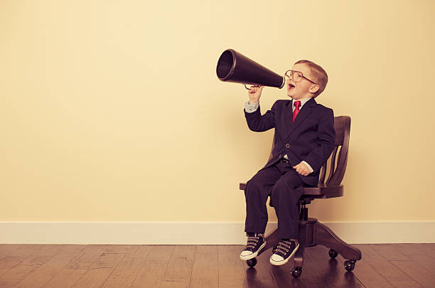 Young Business Boy Sitting in Chair Yelling Through Megaphone This important business message is brought to you by a very smart little businessman. chairperson stock pictures, royalty-free photos & images