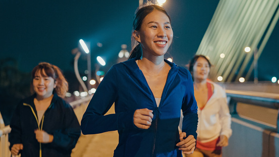 Multiracial team young sporty Asian women wear sports outfits running enjoy cardio through the city streets over bridge at night. Running club and exercise outdoor concept.