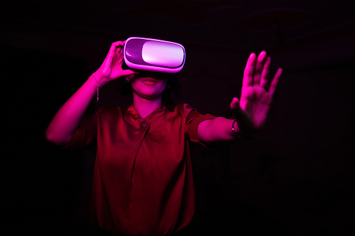 Young indian woman wearing virtual reality goggles headset, asian female have fun with vr box in the dark with colorful pink light, technology and met averse concept.