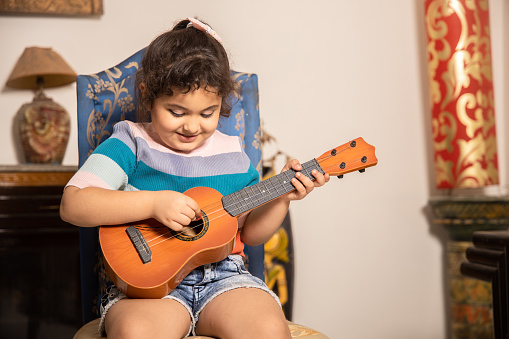 Happy little Indian girl playing acoustic guitar or ukulele at home, learning music.