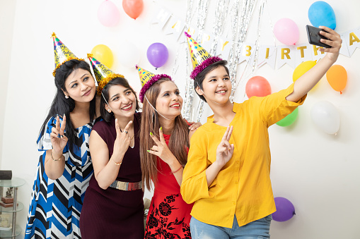 Portrait of smiling young Indian girls making party and celebrating birthday together. Clicking selfie pictures with smartphone