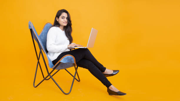 オレンジの黄色い背景に白いtシャツを着た若いインド人女の子が、ノートパソコンを使って椅子に座っている。スタジオショット、コピースペース、コンピューターを使用するアジア人女性 - isolated on white full length lifestyles yellow ストックフォトと画像
