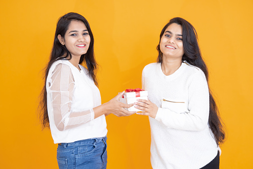 Happy young indian girls wearing white T-shirt exchange gift isolated over yellow orange background. studio shot. Festive celebration. Two happy brunette female friends exchanging presents,
