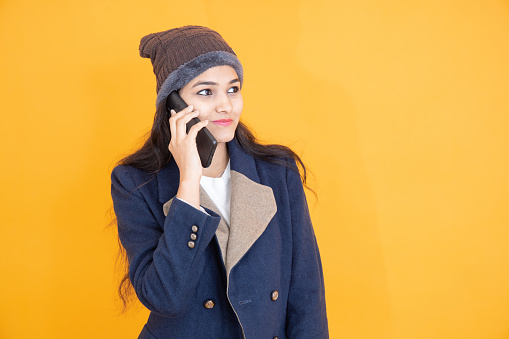 Happy young Indian girl wears blue winter jacket knitted hat talking on mobile phone isolated on orange yellow background studio