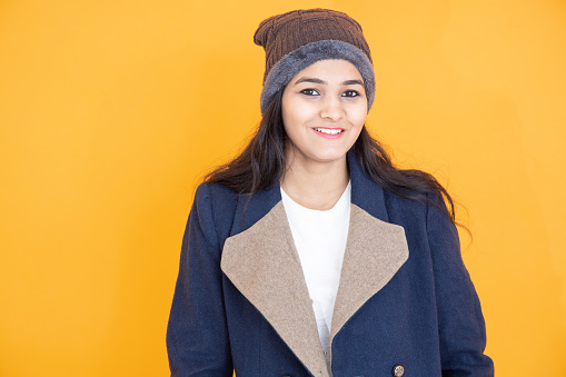 Portrait of happy young indian girl wearing winter cloths smiling isolated over orange yellow background. She is wearing woolen hat and blue jacket or over coat.