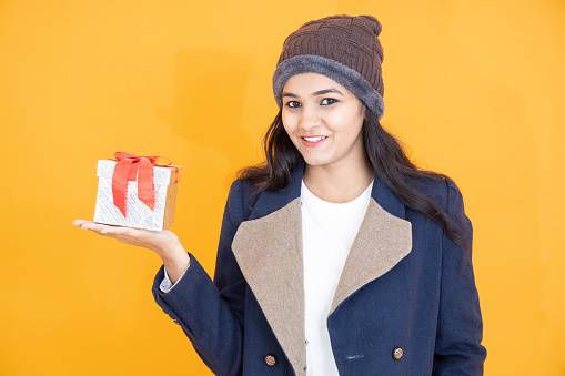 Happy young Indian girl wearing winter cloths holding christmas gift box isolated on orange yellow background, Asian female with knitted hat and blue overcoat jacket.