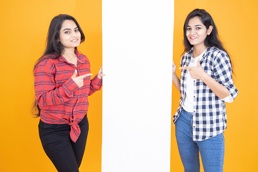 Two Young indian girls standing with blank banner standing to put advertisement isolated over yellow orange studio background. Studio shot, Copy space.