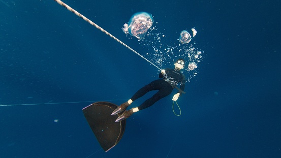 Freediving on the rope in a sea. Male freediver makes bubbles underwater